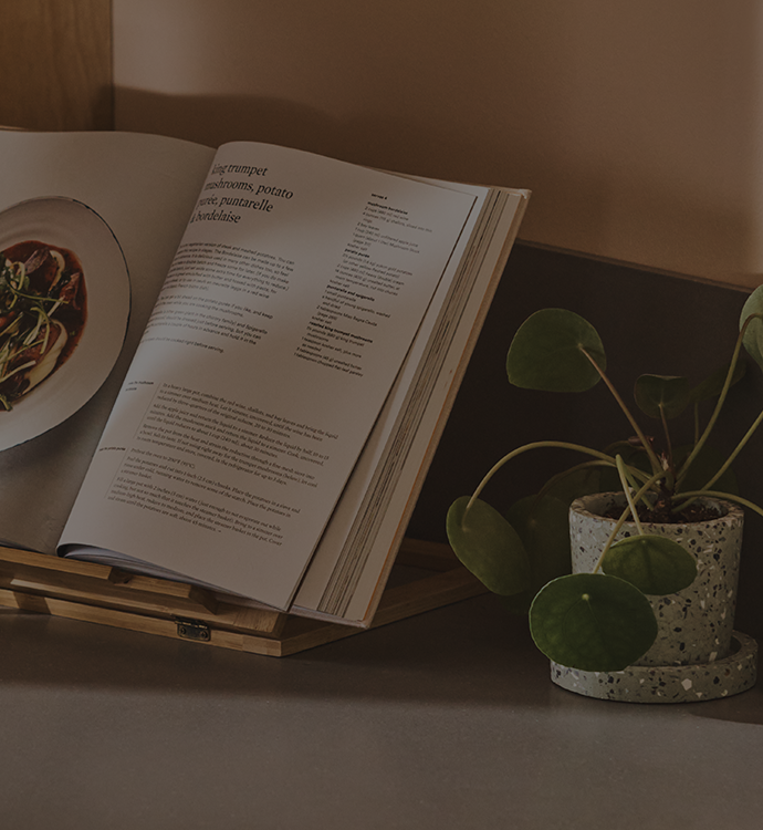 An open cookbook displayed on a wooden stand on a tabletop, with a potted plant beside it and a softly lit interior background.
