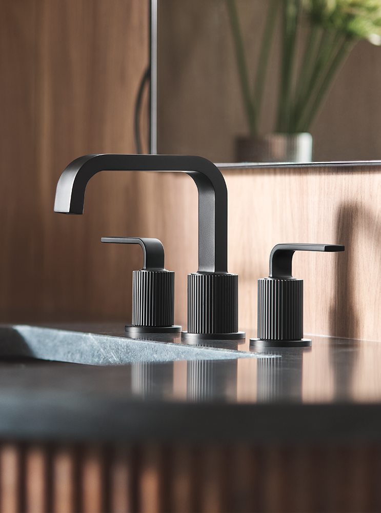 A modern three-hole bathroom faucet set in matte black, installed on a dark stone sink with a wooden backsplash and a mirror in the background.