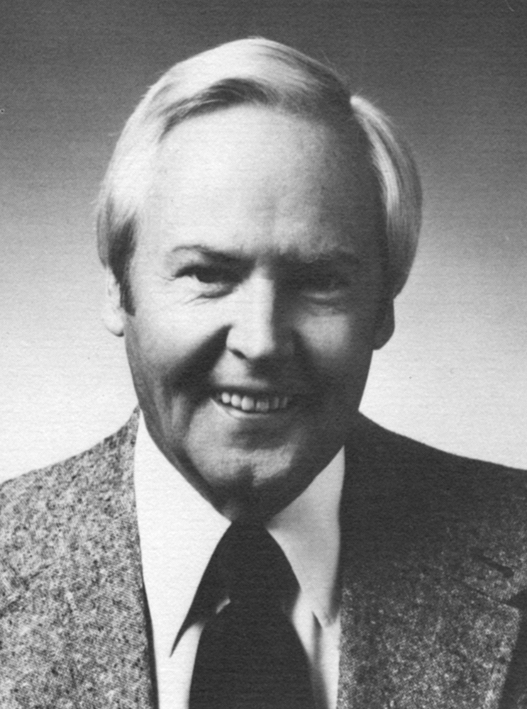 A black and white portrait of an older man with white hair, smiling, wearing a textured tweed jacket, white shirt, and dark tie against a gradient gray background.