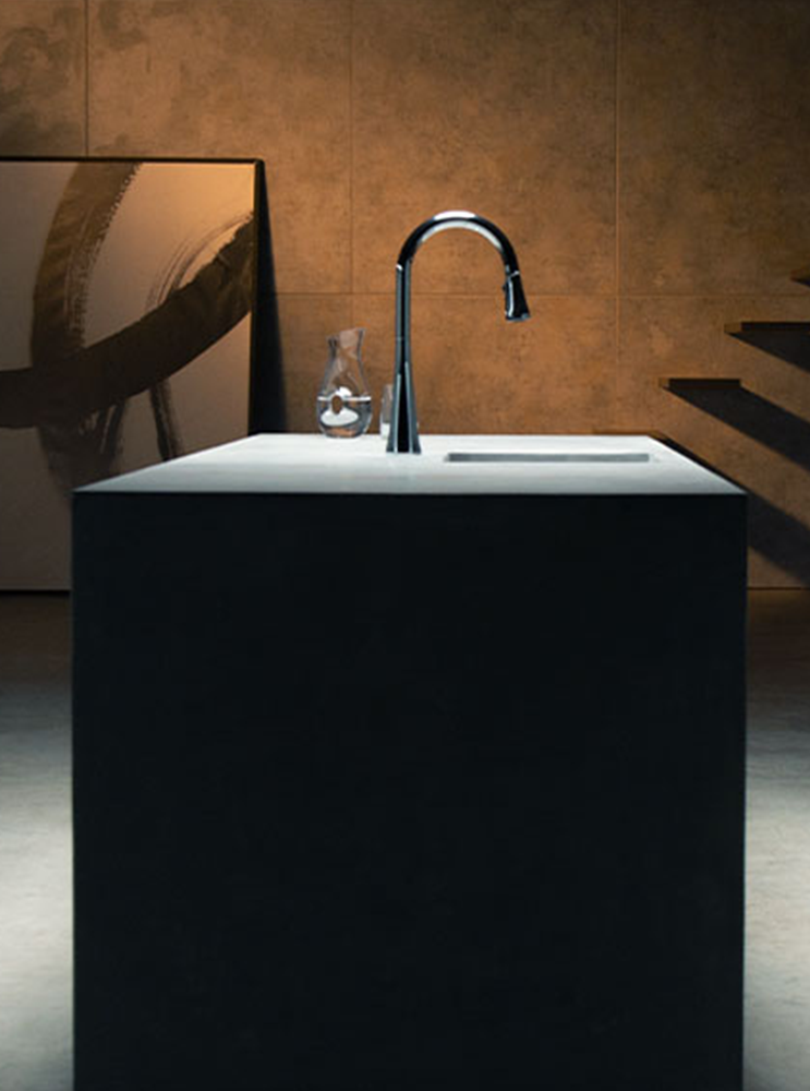 A modern chrome gooseneck faucet mounted on a white countertop, set on a dark pedestal island in a dimly lit room with a textured brown wall and abstract art.