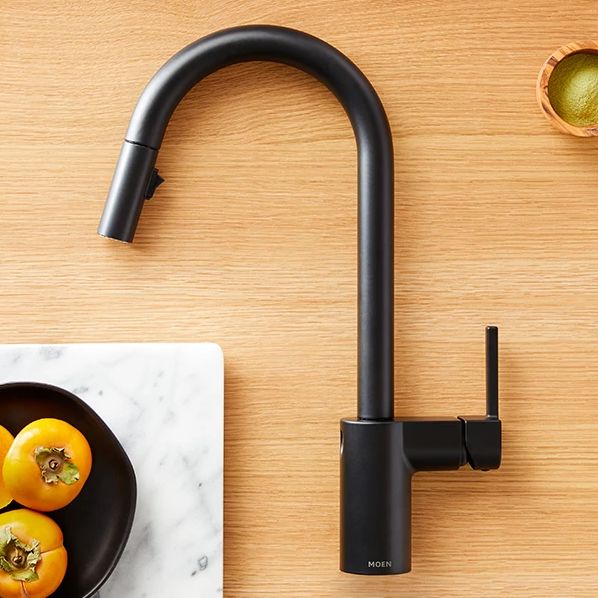 A matte black Moen pull-down kitchen faucet shown from above on a light wood tabletop, with a marble board, yellow fruit, and a small bowl of greenery as styled props.