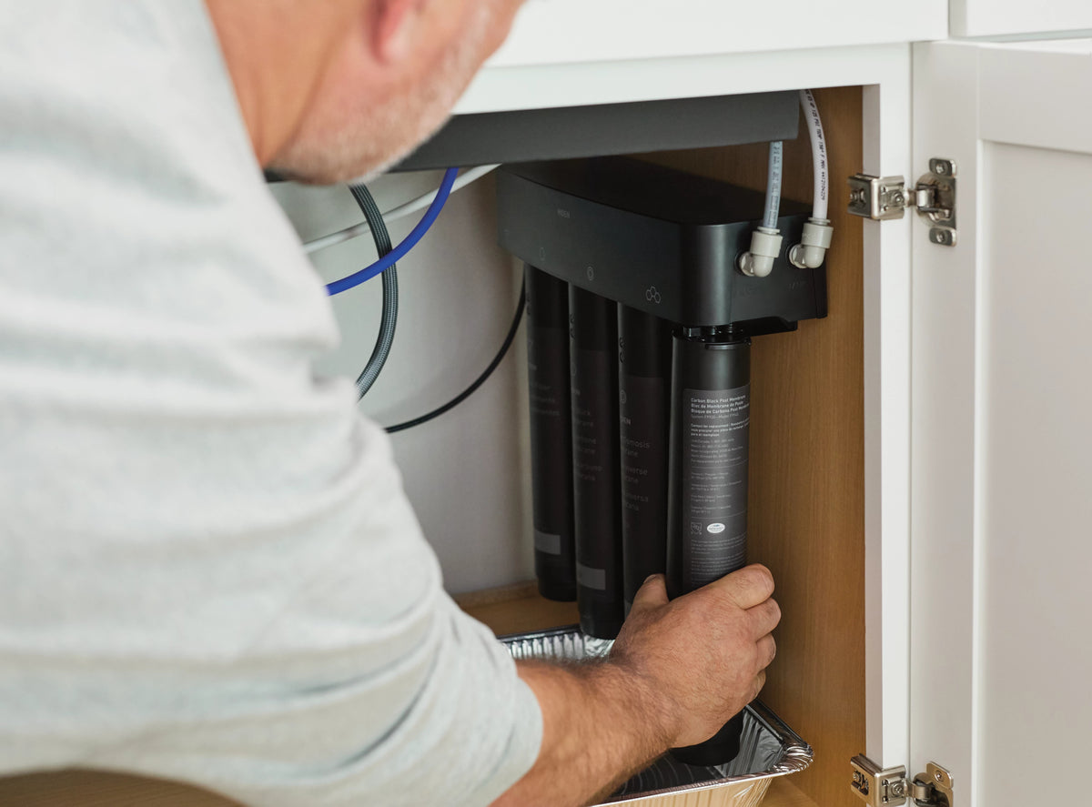 A man in a light gray t-shirt installs a black cylindrical water filter cartridge into a set of three filters under a white kitchen cabinet.