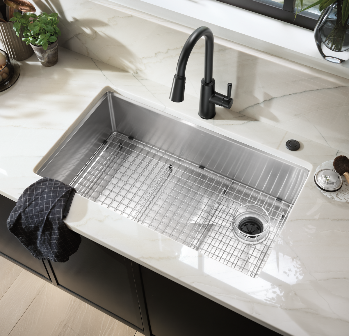 A stainless steel undermount sink with a wire grid drain insert, paired with a matte black gooseneck faucet, set into a white marble kitchen countertop.