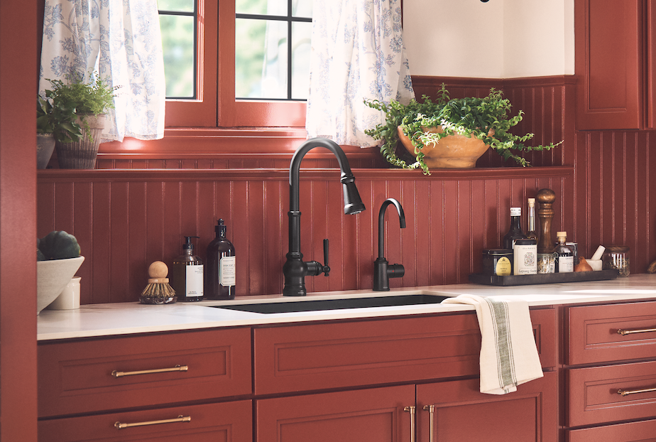 A kitchen scene with red cabinetry, a white countertop, a black sink, and two black faucets. A window with floral curtains and potted plants is visible above the counter.