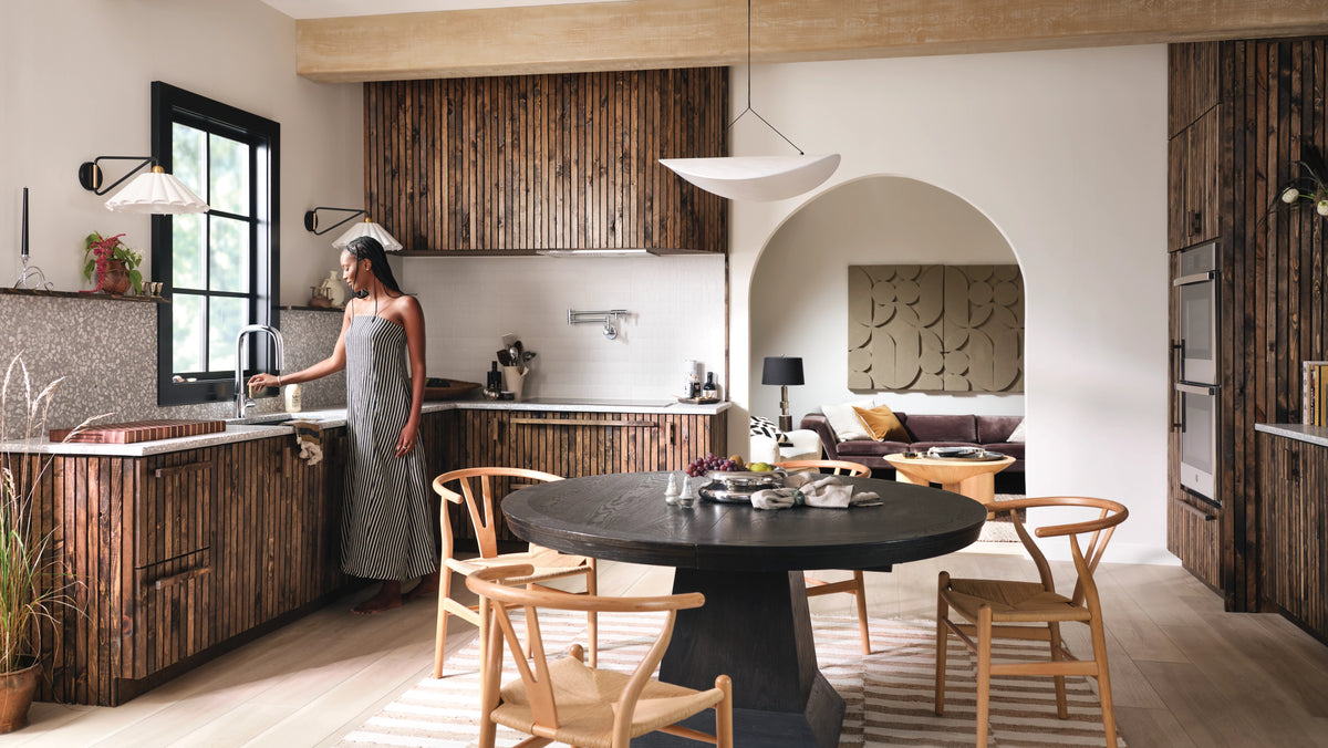 A woman in a sleeveless gray dress stands at a kitchen sink in a modern open-plan kitchen with wood slat cabinets, a round black dining table, and wooden chairs. The room features warm neutral tones, an arched doorway leading to a living area, and a large black pendant light overhead.