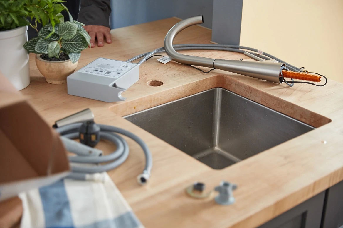 A modern under-counter sink faucet setup installed in a light wood countertop. The stainless steel sink basin is shown with a curved gooseneck faucet, gray hoses, a gray electrical box, and small hardware pieces on the counter.