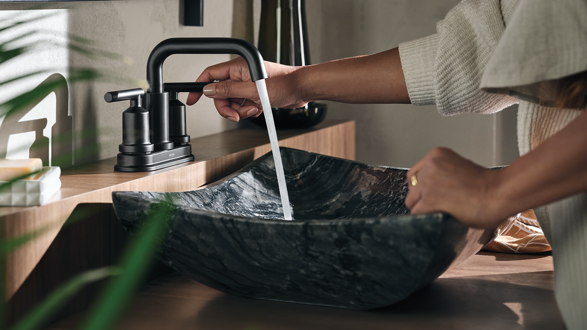 A person washing their hands under a modern matte black faucet in a bathroom setting, with a dark marble vessel sink below.