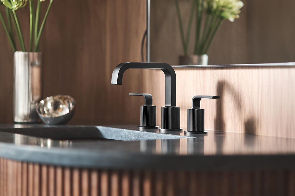 Cambium Two-Handle High Arc Bathroom Faucet, installed on a dark stone sink with a matching black soap dispenser, set against a warm wood-paneled background.