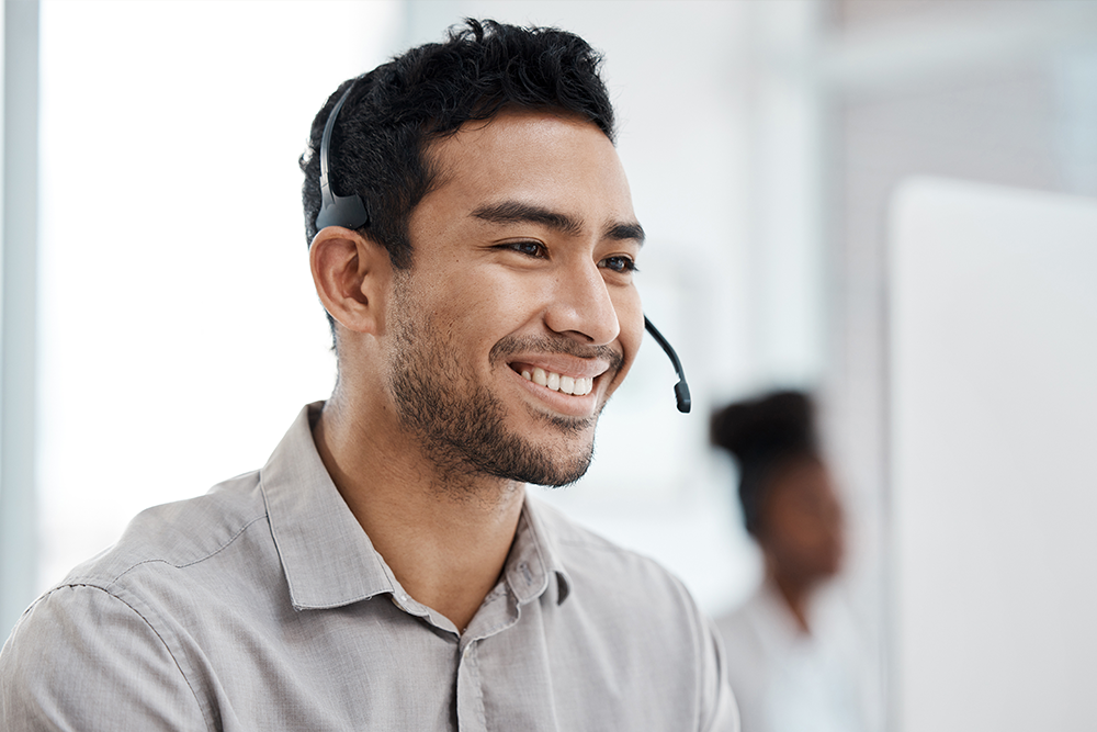 A smiling man wearing a gray button-down shirt and a black headset, photographed in a bright office setting with a blurred colleague in the background.