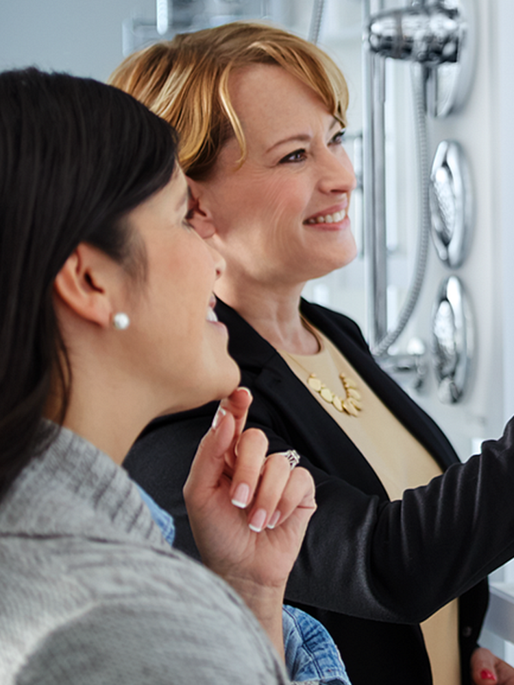 Two women in a showroom looking at wall-mounted chrome shower fixtures. One woman wears a black blazer over a beige top with a gold necklace, and the other is in the foreground wearing a gray top.