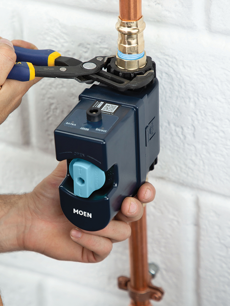 A person's hands using blue and yellow pliers to adjust a copper pipe fitting on a dark blue MOEN water heater control valve, set against a white textured brick wall.