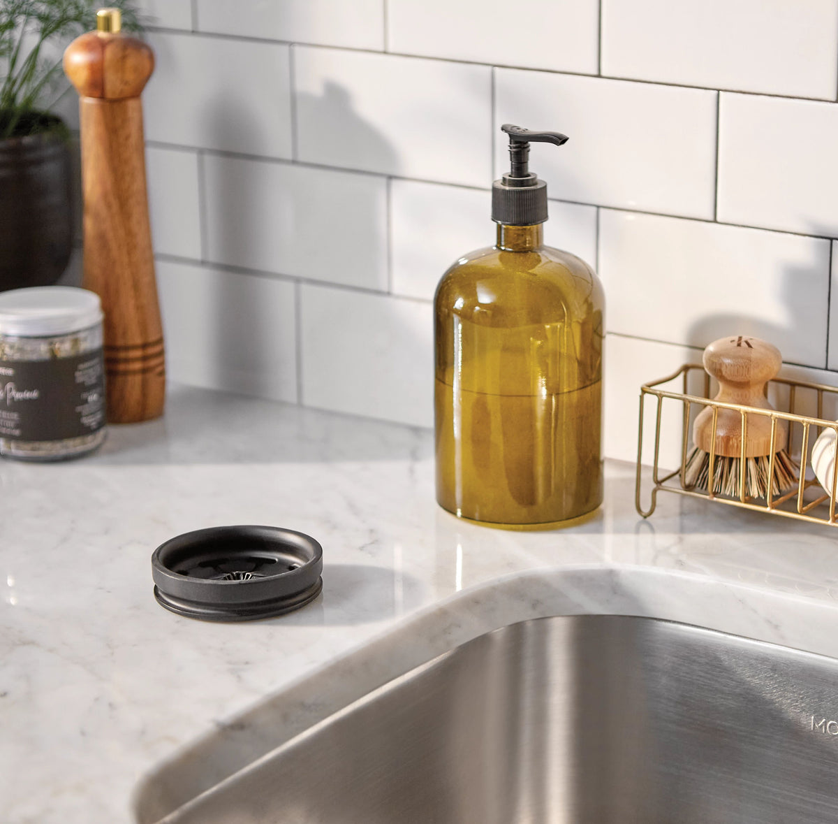 An amber glass soap dispenser with a black pump sits on a white marble kitchen countertop next to a stainless steel sink. A matching black lid is placed in the foreground, with a white subway tile backsplash and kitchen accessories in the background.