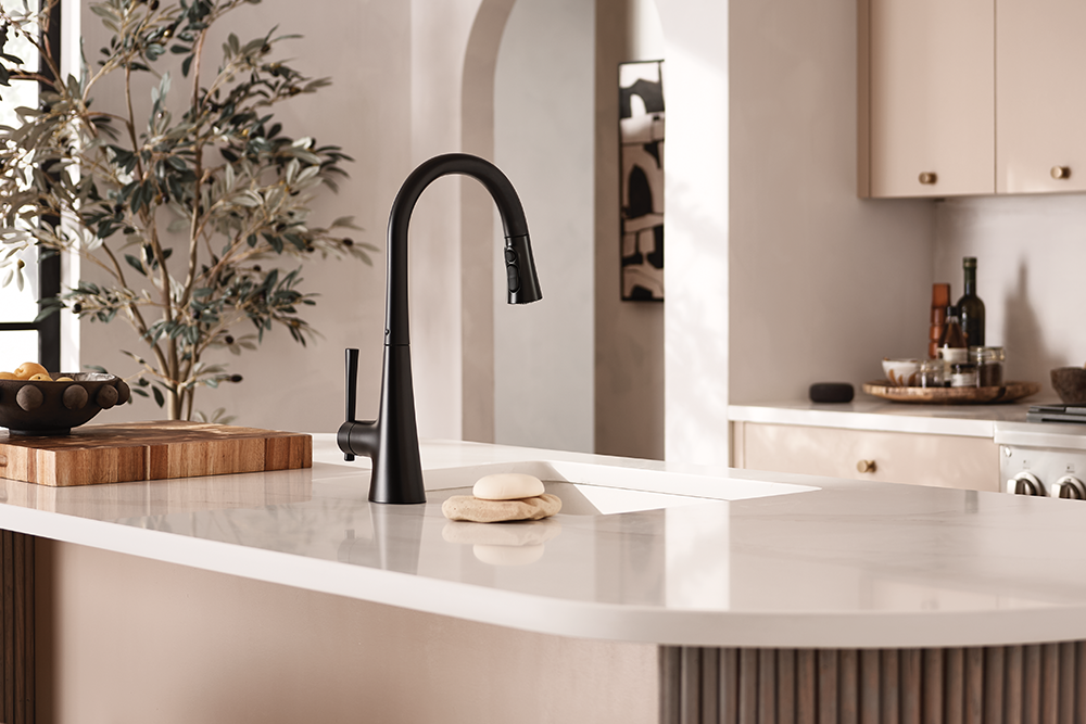 A matte black single-handle kitchen faucet installed on a white countertop in a modern, warm-toned kitchen with beige cabinetry and a decorative plant.