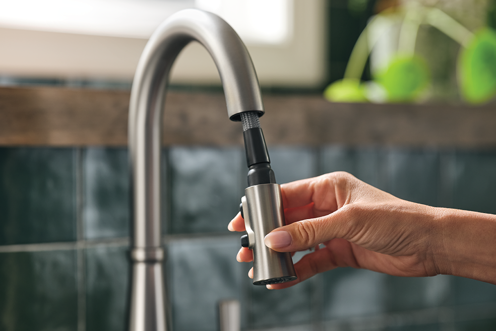 A hand holding a brushed stainless steel and black faucet sprayer attachment under a curved matte stainless steel kitchen faucet in a kitchen setting with dark tiled backsplash.
