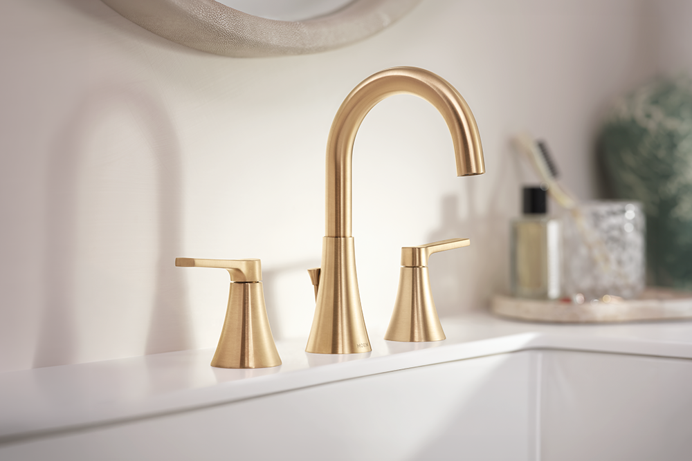 Jase Two-Handle High Arc Bathroom Faucet set in brushed gold, featuring a tall curved spout and two separate lever handles, installed on a white sink with a softly blurred bathroom background.