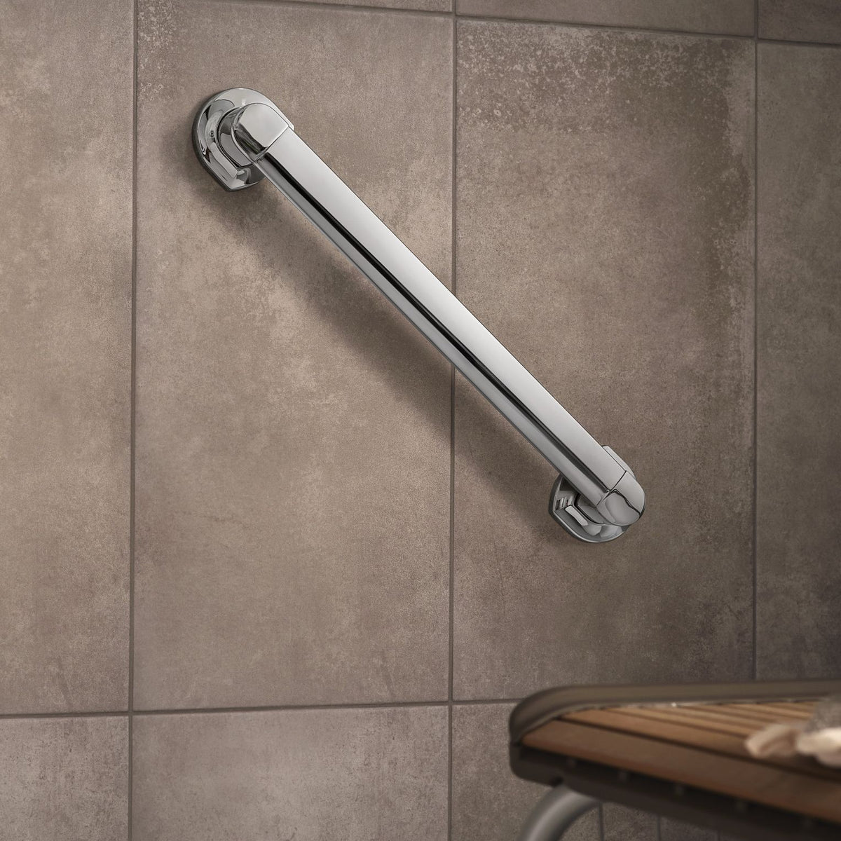 A polished chrome bathroom grab bar mounted on a textured taupe tile wall, with part of a wooden bench visible in the lower right corner.