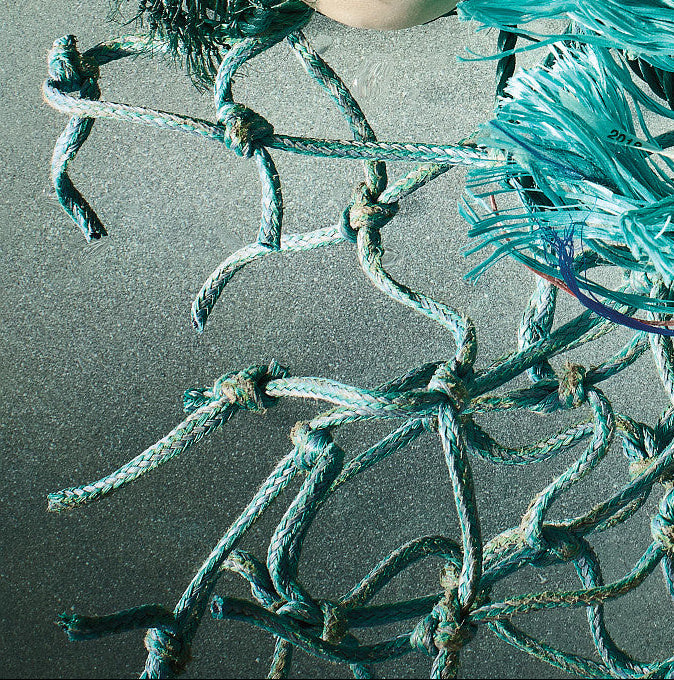 A close-up of tangled teal and turquoise ropes with knots, alongside small seashells and pebbles on a textured gray surface.