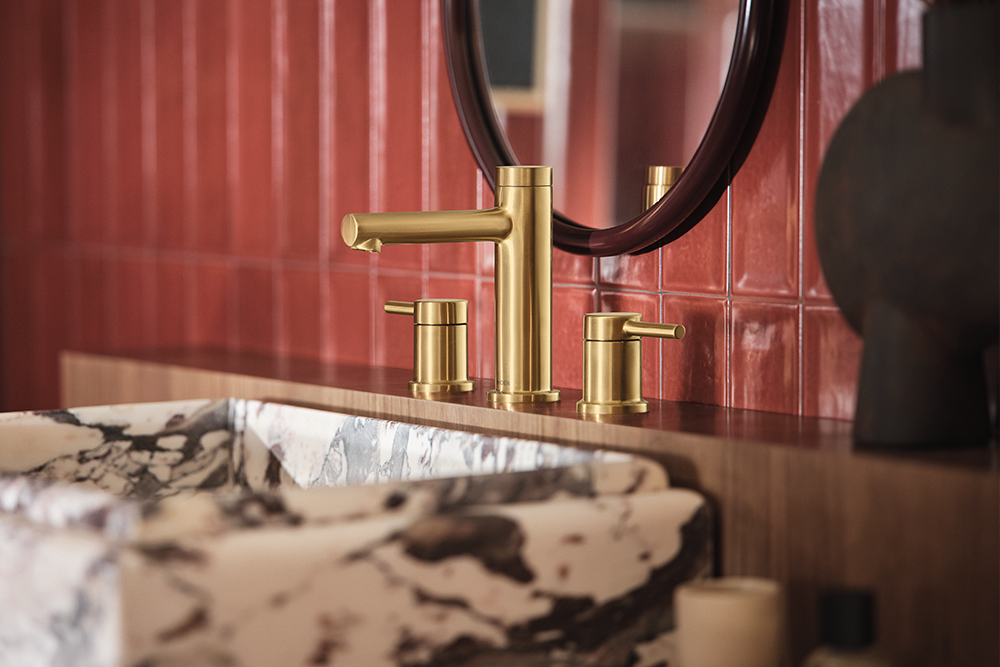 A close-up of a modern bathroom faucet set in brushed gold, mounted on a wooden countertop with a black and white marble sink basin and a red tiled wall background.
