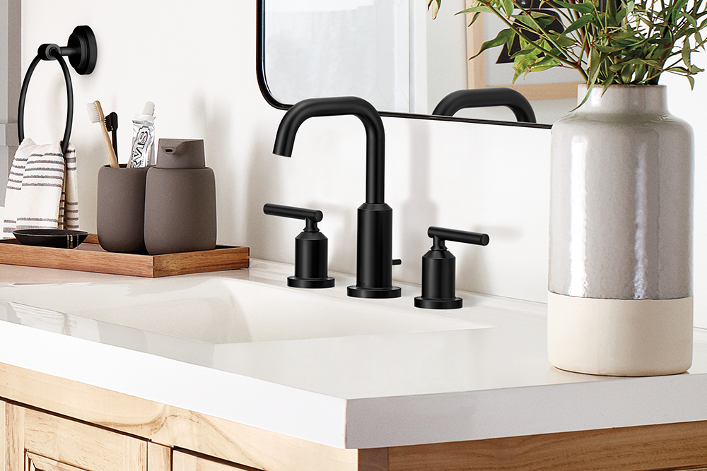 A modern three-hole bathroom faucet set in matte black, installed on a white countertop with a light wood vanity. The scene includes a matching black wall-mounted towel ring, gray ceramic tooth holders, a large gray and cream vase with greenery, and a mirror reflecting the setup.