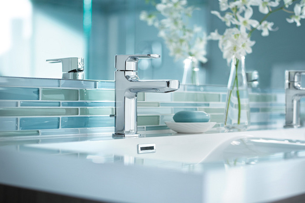 A modern chrome single-handle bathroom faucet installed on a white sink, with a blue-tiled backsplash, a soap dish with a bar of soap, and white flowers in the background.