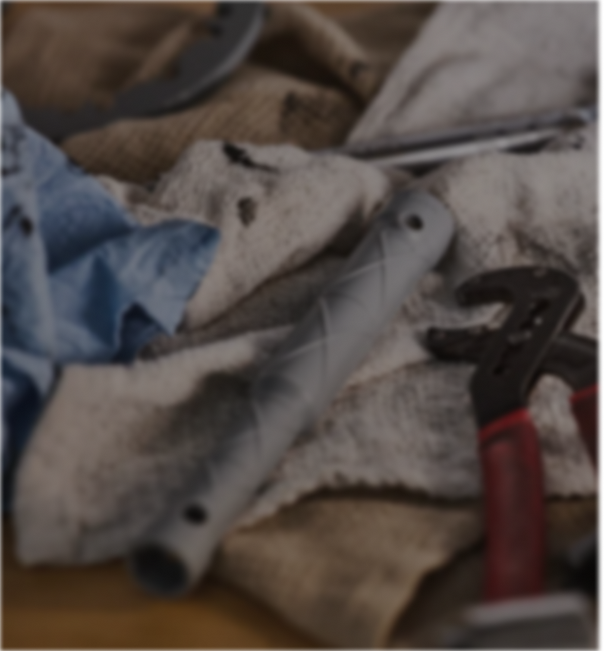 A close-up, slightly blurred photo of workshop tools and worn fabric, including a gray handled tool, a red-handled wrench, and cloth pieces on a workbench.