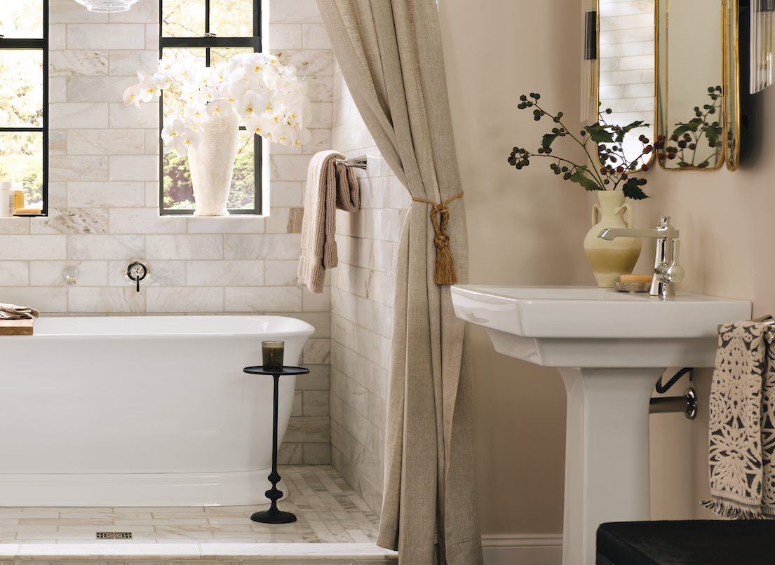 A luxury bathroom scene featuring a white freestanding clawfoot bathtub set in a tiled corner with beige curtains, a white pedestal sink, and decorative accents.