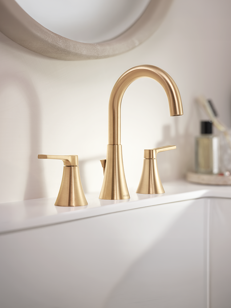A modern three-hole bathroom faucet set in brushed gold, featuring a tall curved spout and two matching lever handles, installed on a white sink countertop with a softly blurred bathroom background.