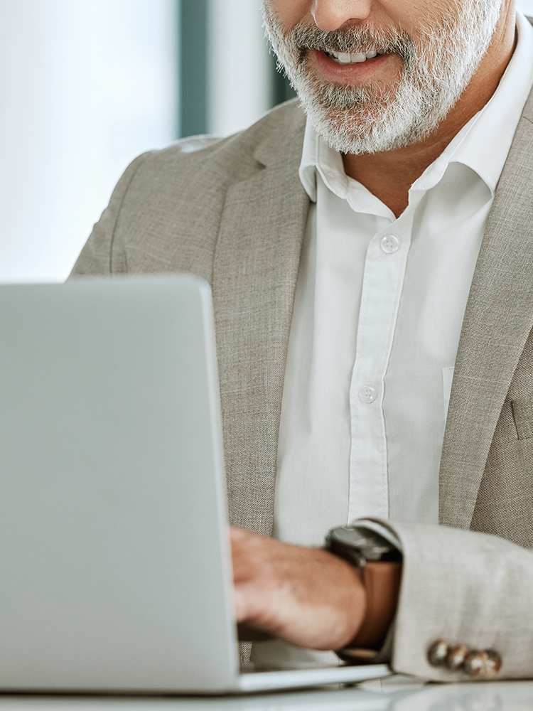 A close-up of a man with a gray beard wearing a beige blazer and white shirt, typing on a silver laptop in a bright office setting.