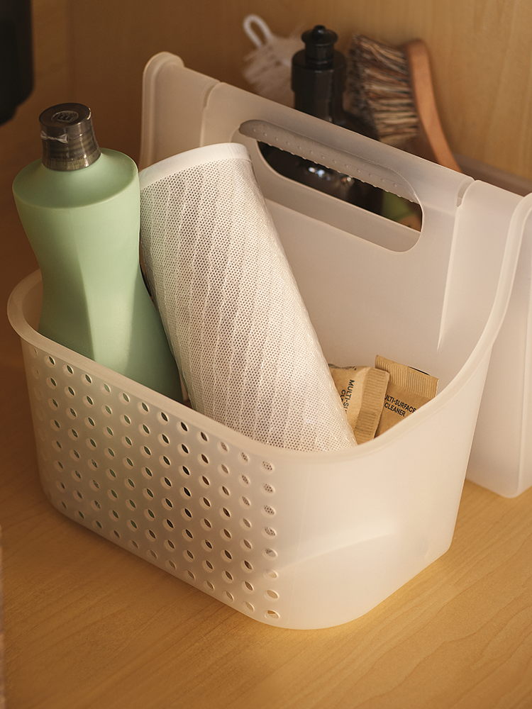 A white plastic organizer basket with a perforated front and integrated handle, holding a mint green bottle, a white mesh bag, and small brown packets on a wooden surface.