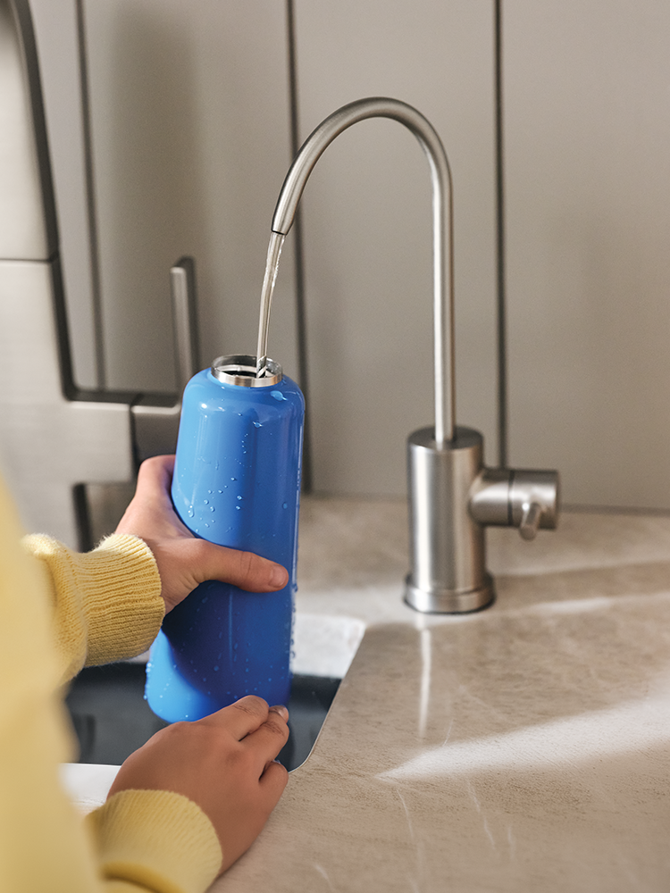 A person holding a blue water filter canister under a stainless steel gooseneck faucet in a modern kitchen setting.