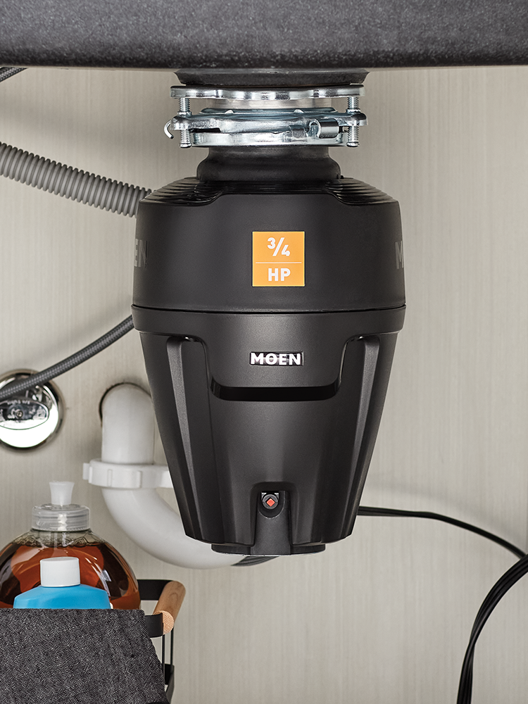 A Moen 3/4 HP commercial-grade undercounter garbage disposal mounted on a beige cabinet wall, with a black sink deck above and plumbing visible.