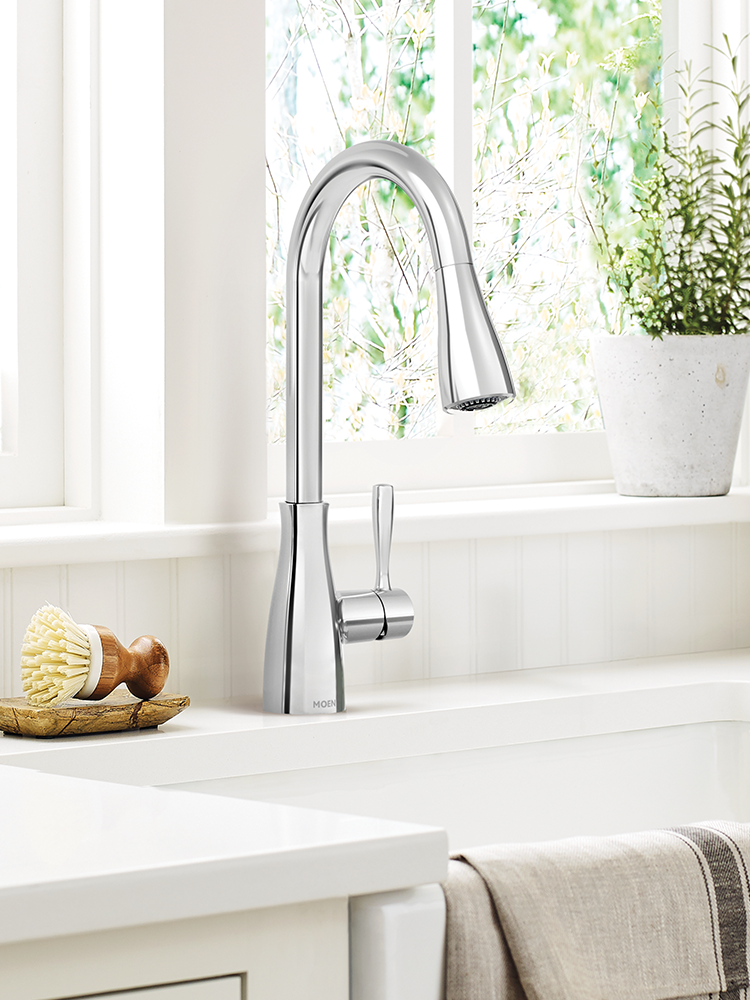A modern polished chrome pull-down kitchen faucet installed on a white sink countertop in a bright kitchen setting with a window, plant, and soap dish visible.