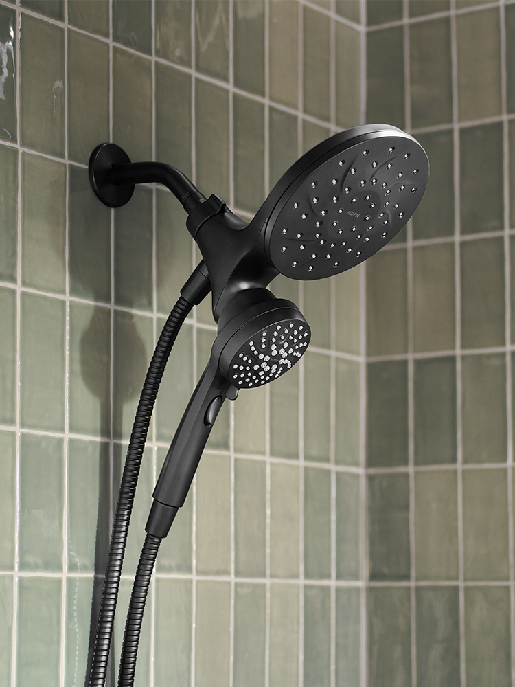 A matte black dual-head shower system with a wall-mounted round shower head, flexible hose, and handheld shower attachment, shown installed against a green tiled bathroom wall.