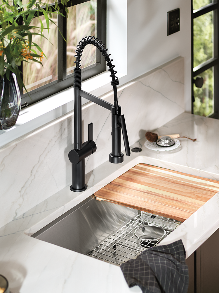 A modern kitchen sink setup featuring a matte black gooseneck faucet mounted on a white marble countertop with a wooden cutting board and stainless steel sink.