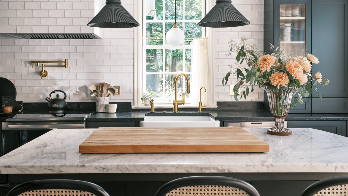 A modern kitchen scene with a large wooden cutting board centered on a marble island countertop. The kitchen features dark charcoal cabinets, white subway tile backsplash, brass fixtures, black pendant lights, and a vase of peach flowers on the counter.