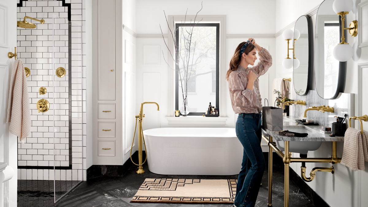 A woman stands in a bright, modern bathroom with white walls, a freestanding bathtub, a walk-in shower with white subway tile and gold fixtures, and a marble vanity with gold hardware. She is wearing a pinkish-beige textured top and blue jeans.