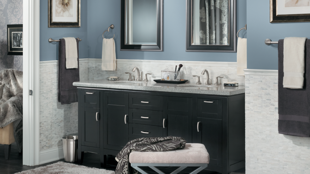 A modern bathroom vanity setup with a black double-sink cabinet, white marble countertop, framed mirrors, blue-gray walls, and matching towels hanging on wall racks.