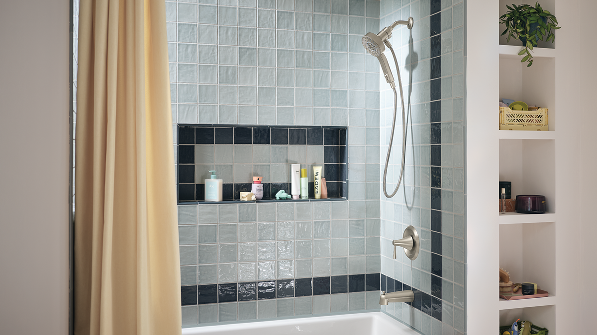 A modern bathroom shower area with glossy light blue-gray square tiles, a built-in dark navy tile niche holding toiletries, a beige shower curtain, and white open shelving on the right.