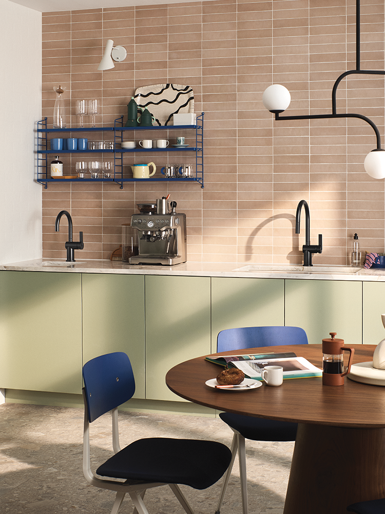 A modern kitchen interior with beige rectangular wall tiles, sage green lower cabinets, black faucets, blue wire shelving, a round wooden dining table, and blue-and-black chairs. No person or animal is present, but this is still a real environment rather than a clean isolated product shot, so subject_type is Lifestyle.