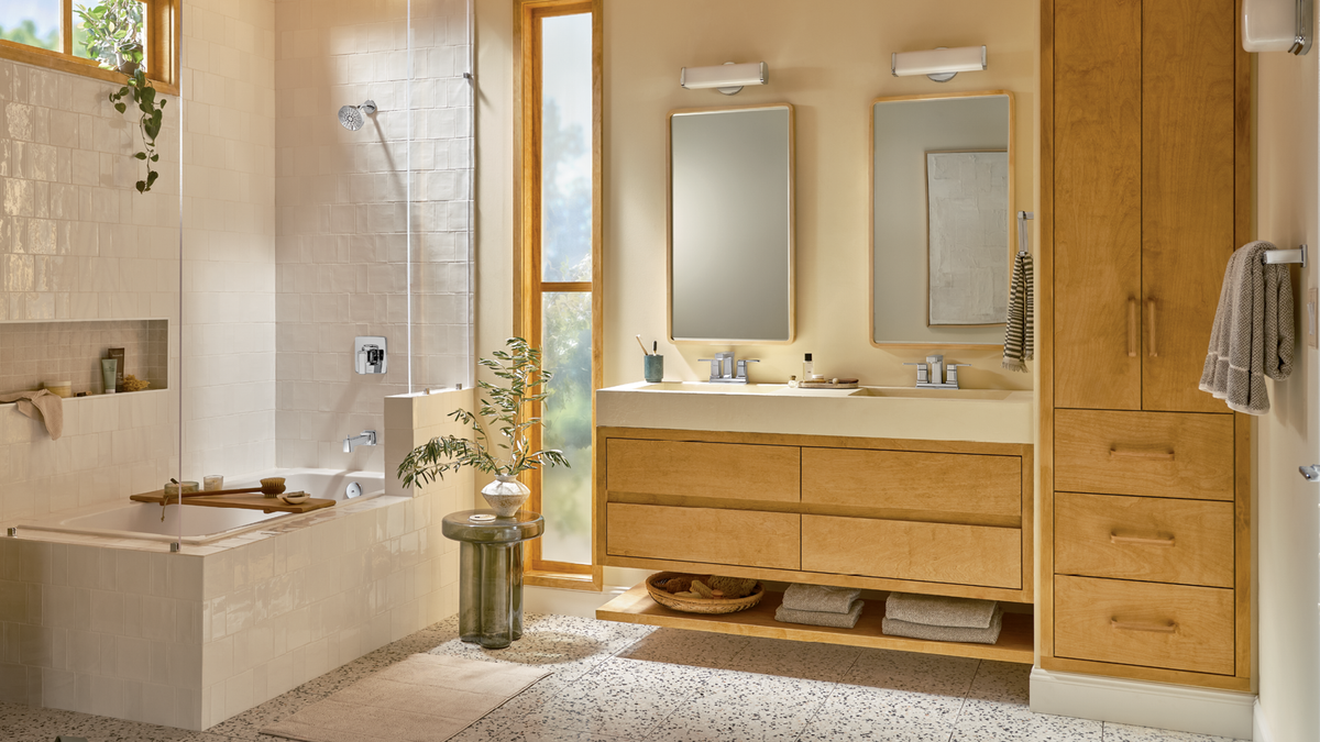 A modern bathroom interior featuring a light wood double-sink vanity with mirrors, a freestanding bathtub, a tall matching wood cabinet, and warm beige walls and flooring.
