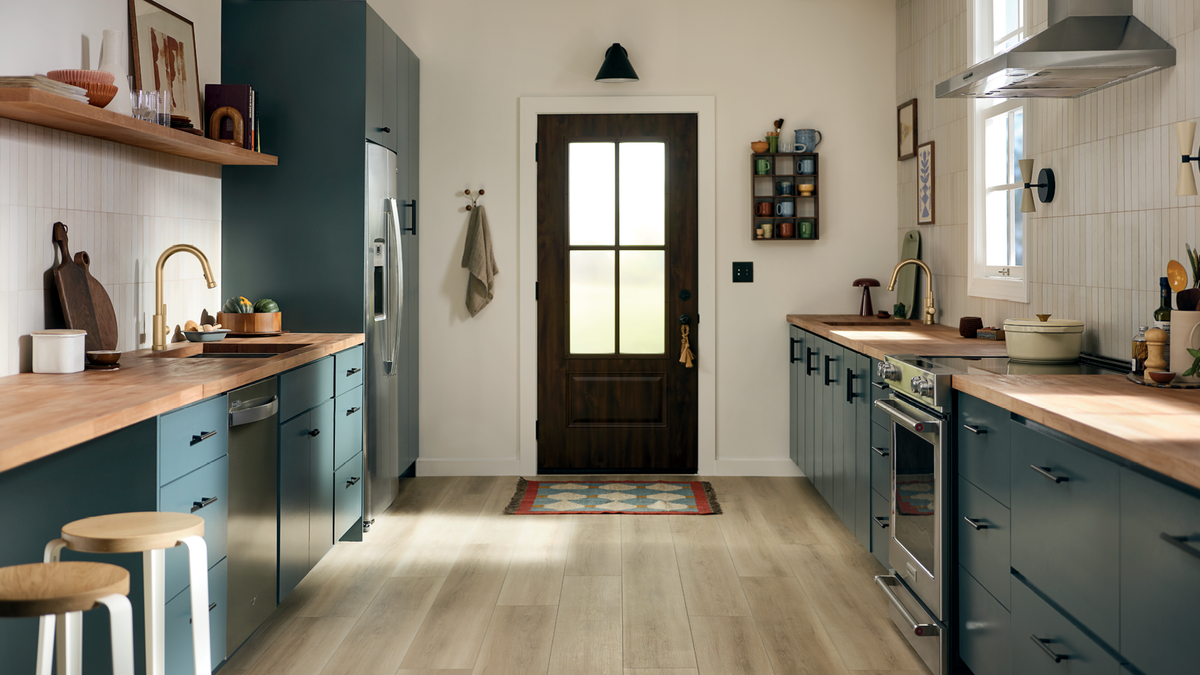 A modern kitchen interior with dark teal cabinets, light wood countertops, white subway tile backsplash, stainless steel appliances, and a dark wood front door at the back with a small multicolor rug in front.