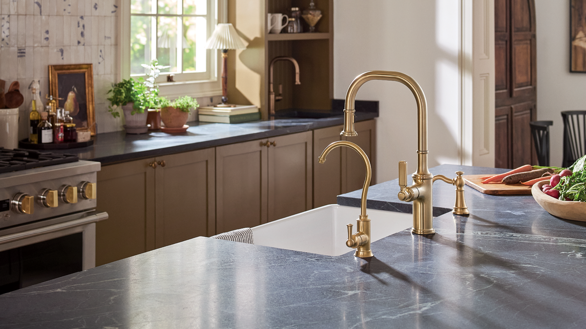 A modern kitchen scene featuring a tall, elegant brushed brass faucet with a matching side lever and a smaller auxiliary spout, installed on a dark stone countertop.