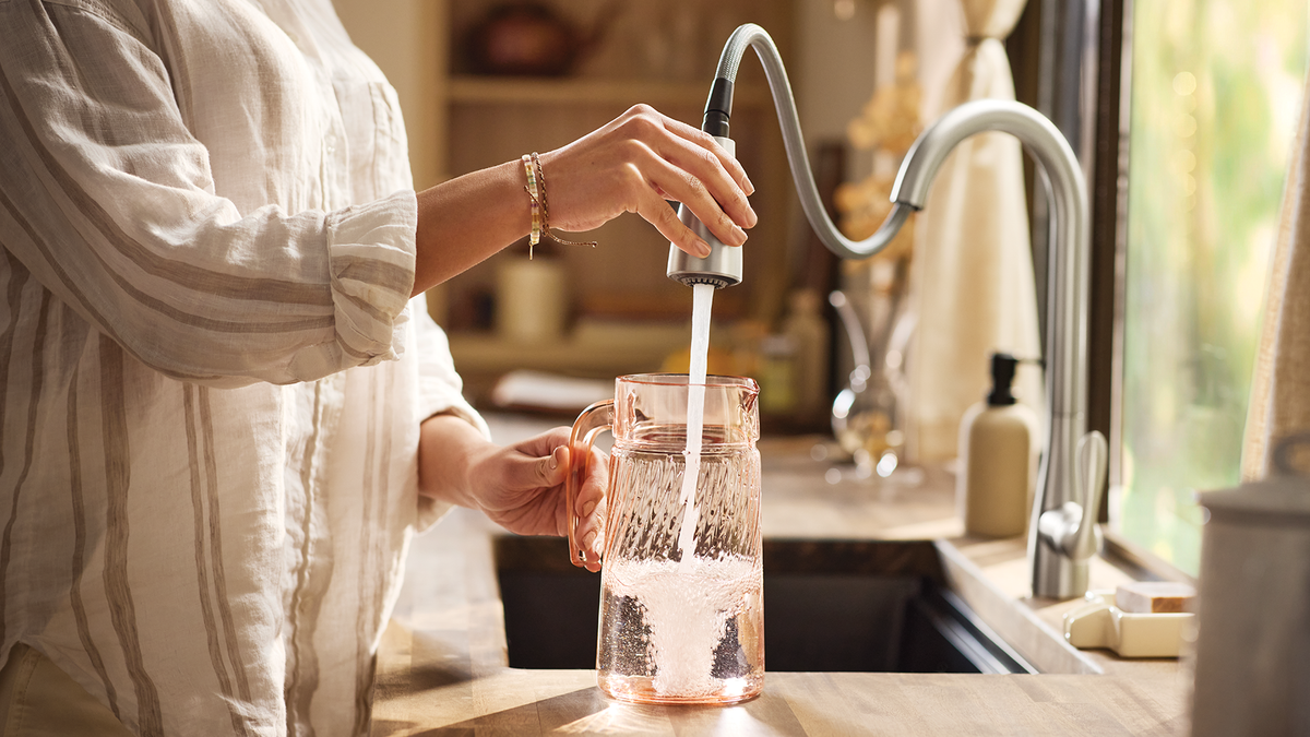 A person in a white striped shirt fills a clear glass pitcher with water from a modern curved faucet in a warm, sunlit kitchen setting.