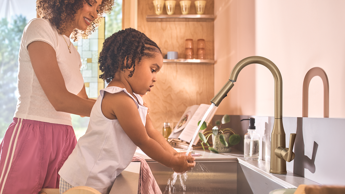 A woman and a young girl wash hands together at a kitchen sink with a modern matte brass faucet. The scene is set in a warm, well-lit kitchen with wooden shelves and neutral tones.
