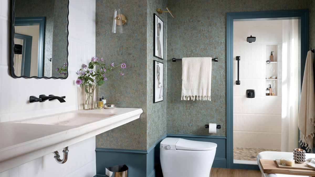 A styled bathroom interior with blue-green patterned wallpaper, blue trim, a white vanity, a white toilet, a mirror, framed wall art, a towel, and an open shower door revealing a bright shower area.