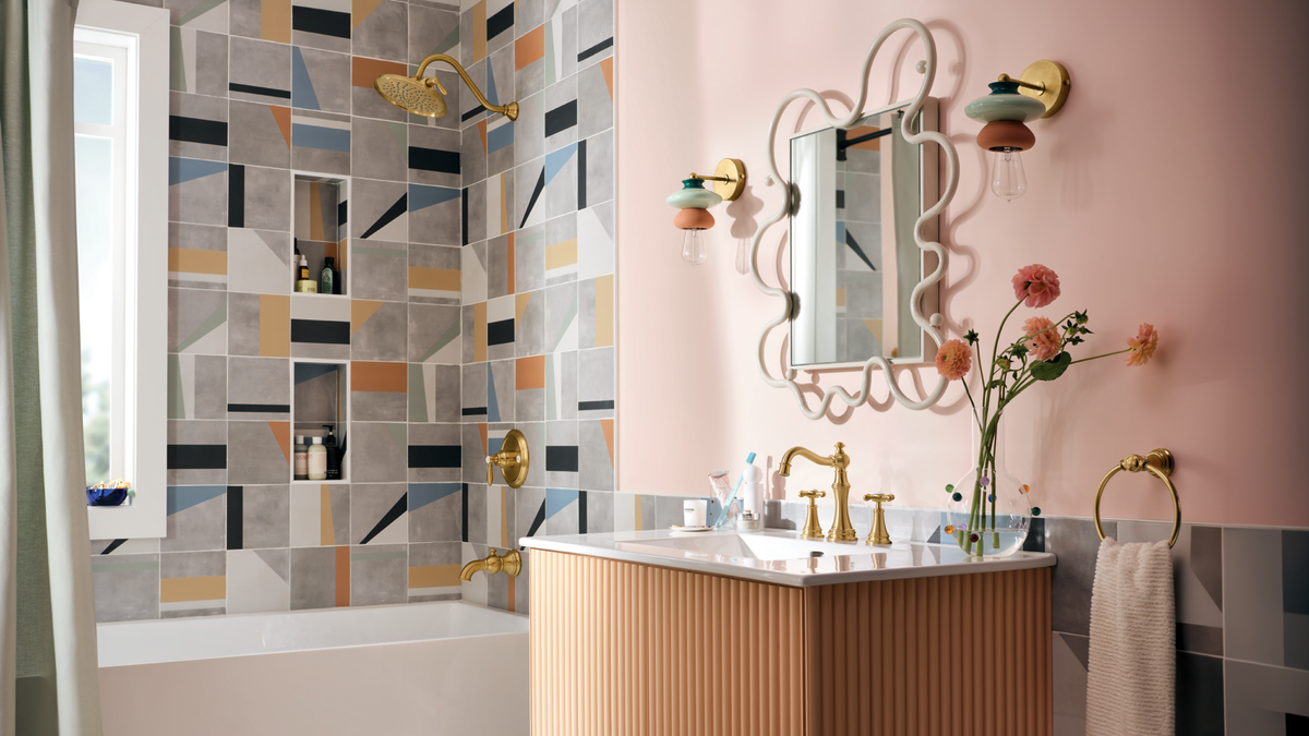 A styled bathroom interior featuring a bathtub with a geometric mosaic tile wall in gray, blue, orange, and beige, a pink wall, a decorative white mirror, gold fixtures, and a vanity with a marble top.