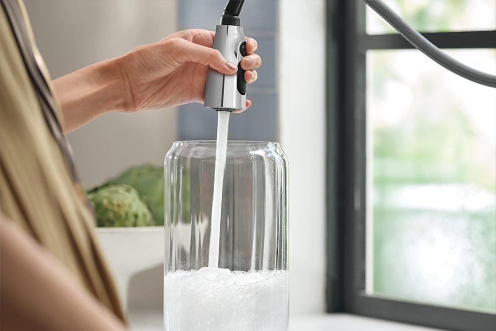 A person's hand holds a silver and black pull-down faucet sprayer above a clear glass water jug, with water flowing into it. The scene is set in a bright kitchen or bathroom next to a window.
