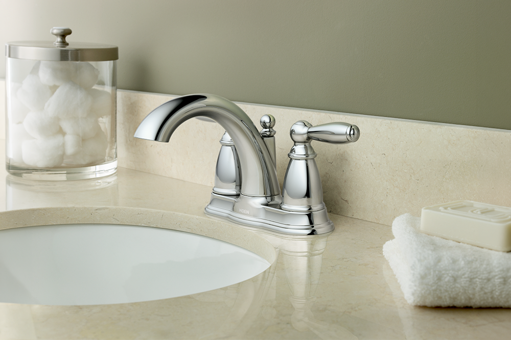 A polished chrome widespread bathroom faucet installed on a beige marble countertop with an undermount sink, next to a glass jar of cotton pads and a folded white towel with soap.