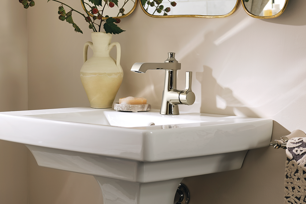 A modern chrome single-handle bathroom faucet installed on a white ceramic pedestal sink, with a cream vase and soap dish on the sink deck, set against a beige wall with decorative mirrors.