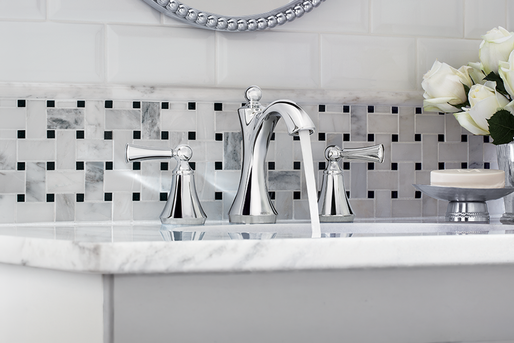 A polished chrome three-hole bathroom faucet with a high-arc spout and two separate lever handles, installed on a white marble sink. The background features a white subway tile wall with a black and white marble mosaic tile backsplash, and a vase of white roses sits on the right.